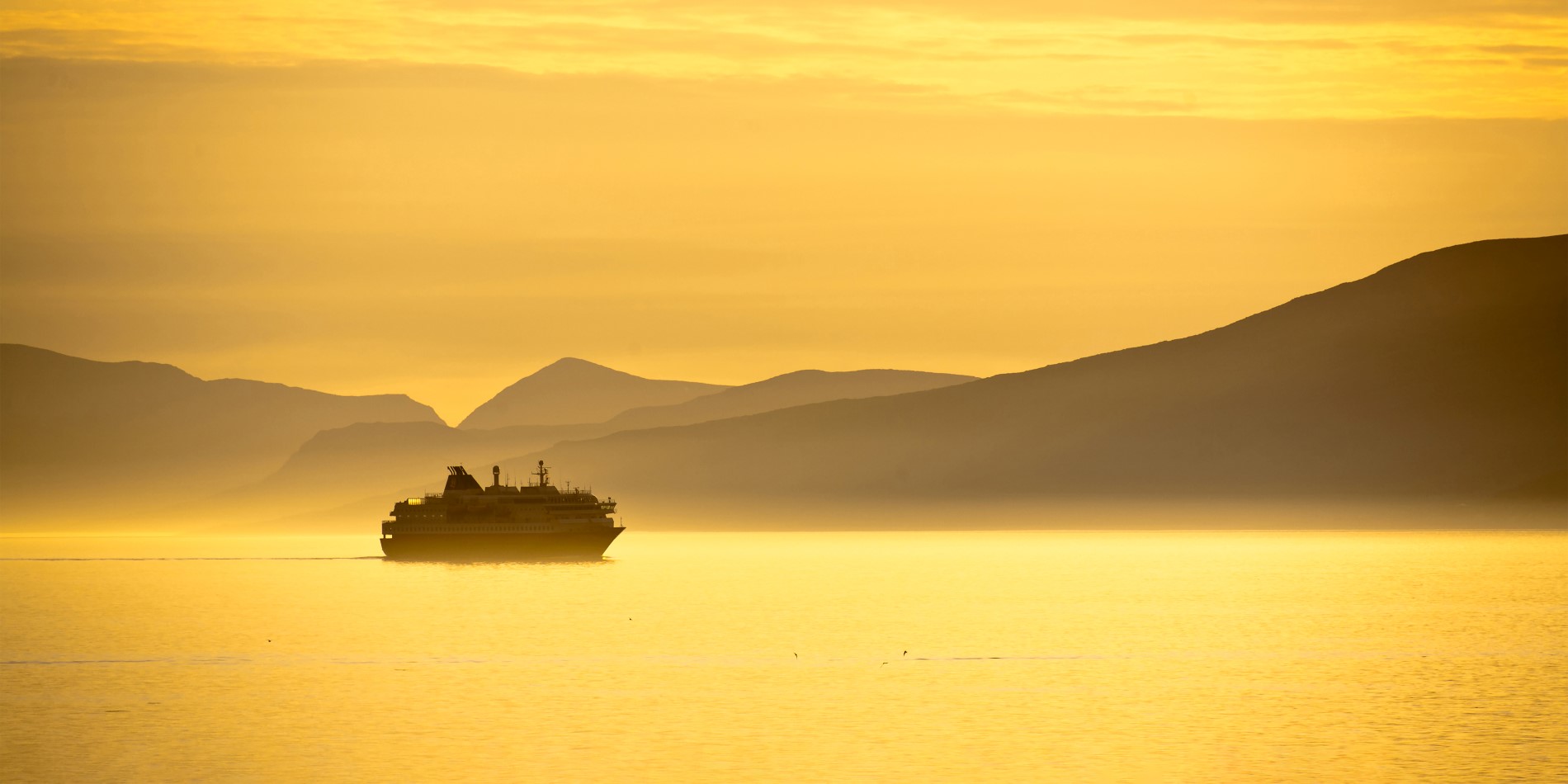 Un navire de Hurtigruten sous le Soleil de Minuit, avec le ciel et l’eau renvoyant une lumière jaune éclatante 