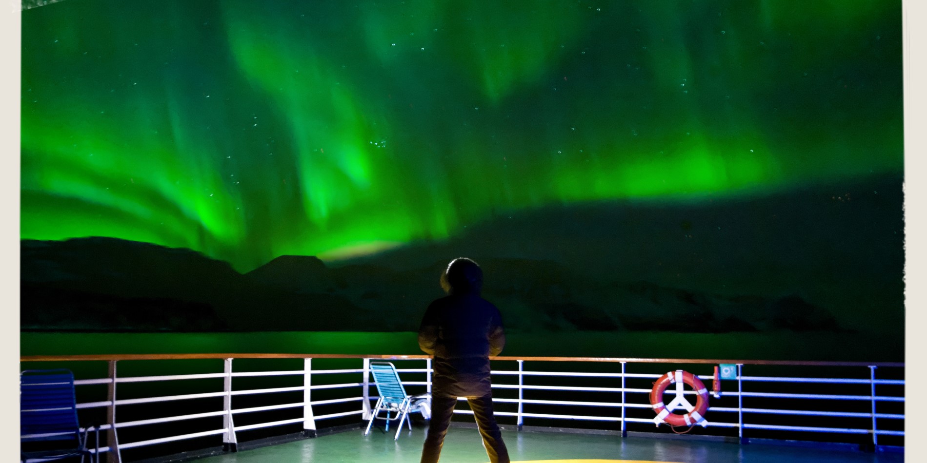 Homme se tenant debout sur le pont de l’Express Côtier de Norvège de Hurtigruten. Au-dessus de lui, le ciel est illuminé de vert ; les Aurores Boréales dansent